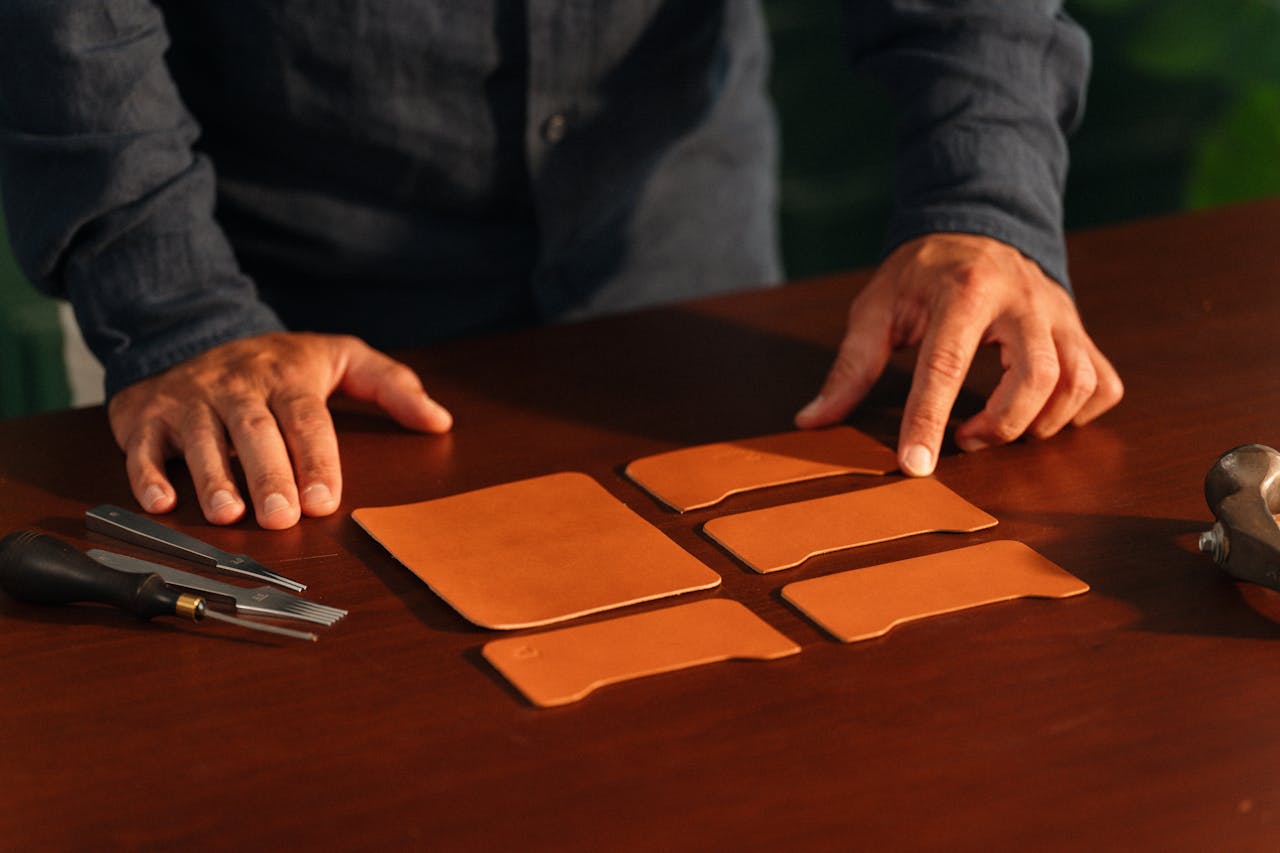 Hands of a craftsman meticulously arranging leather pieces with tools on a wooden table.